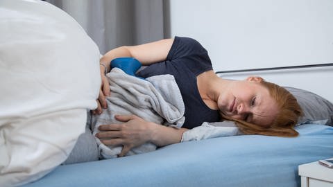 Junge Frau liegt mit Wärmflasche im Bett; sie hat starke Regelschmerzen: Zermürbende Schmerzen, Ungewissheit, Unverständnis – Endometriose führt häufig zu starkem psychischen Leid