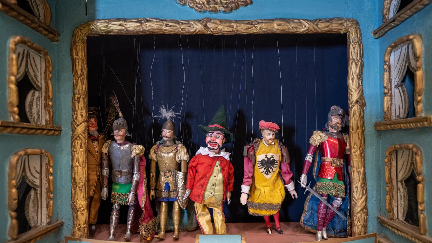Ein historisches Puppentheater von circa 1870 steht im Münchner Marionettentheater neben der Bühne: Marionetten erscheinen auf der Bühne u. a. deshalb so groß, weil die Requisiten – also die Gegenstände, mit denen sie zu tun haben, wie Stühle, Tische oder Türen – im Verhältnis etwas kleiner gestaltet sind.