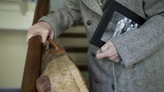 Seniorin mit Koffer und Familienfoto unter dem Arm steigt eine Treppe hinunter: Orientierungsstörungen gehören auf mit zur Demenz. Die sind am Anfang zwar nicht so deutlich, nehmen aber zu.