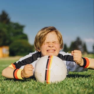 Junge im Trikot der deutschen Fußballmannschaft liegt im Gras, vor sich einen Ball, und drückt die Daumen: Den Ausdruck kannten offenbar schon die alten Römer, für die das Daumendrücken zu tun hatte mit "jemandem Glück wünschen", "jemanden in Gedanken unterstützen". 