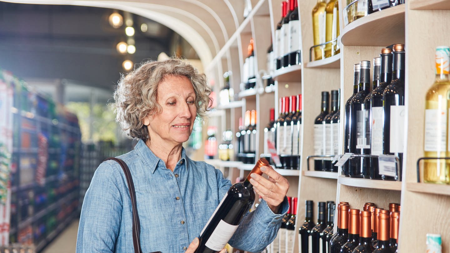 Kundin im Supermarkt vor dem Weinregal mit einer Flasche Rotwein in der Hand: Die meisten Weine aus dem Supermarkt sollte man so jung wie möglich trinken. Wenn man beim Einkauf zum neuesten Jahrgang greift, macht man nichts falsch.