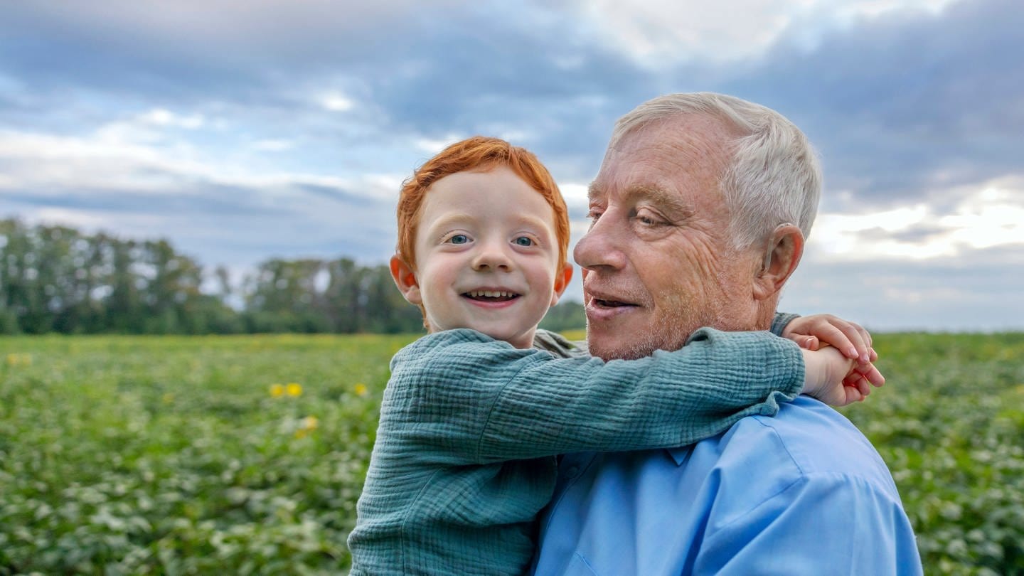 Ein älterer Mann steht am Rand eines Feldes und hält seinen kleinen Enkel auf dem Arm: Für Kinder dauert ein Jahr eine gefühlte Ewigkeit, im Alter geht die Zeit im Nu vorbei. Dieses Phänomen ist inzwischen gut belegt und lässt sich psychologisch erklären.