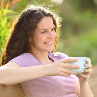 Junge Frau sitzt lächelnd mit einer Kaffeetasse auf einer Bank im Grünen: Der Mensch soll täglich möglichst anderthalb bis zwei Liter Wasser pro Tag trinken, bei Hitze entsprechend mehr. Ist Kaffee hierfür geeignet?