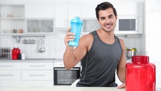 Junger Mann steht im Sportdress in einer Küche und hält eine Trinkflasche, vor ihm eine Dose mit Proteinpulver: Die Deutsche Gesellschaft für Ernährung (DGE) empfiehlt, dass wir am Tag 0,8 Gramm Protein pro Kilogramm Körpergewicht zu uns nehmen sollen. Wenn wir aber viel Sport machen, darf es auch etwas mehr sein.