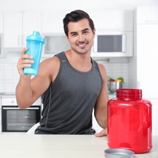 Junger Mann steht im Sportdress in einer Küche und hält eine Trinkflasche, vor ihm eine Dose mit Proteinpulver: Die Deutsche Gesellschaft für Ernährung (DGE) empfiehlt, dass wir am Tag 0,8 Gramm Protein pro Kilogramm Körpergewicht zu uns nehmen sollen. Wenn wir aber viel Sport machen, darf es auch etwas mehr sein.