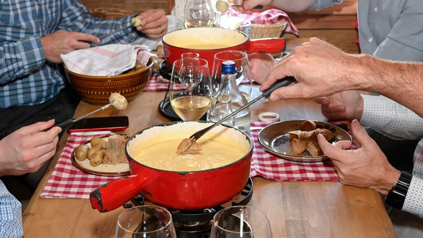 Vier Leute sitzen an einem Tisch und haben zwei rote Töpfe mit Käsefondue vor sich: Käse besteht im Wesentlichen aus Kaseinen; die haben eine gewisse Denaturierungstemperatur. Die Kaseine denaturieren, wenn es ihnen im Fonduetopf zu warm wird. Dann trennt sich das Fett von den Eiweißen. Mit einem Trick lässt sich das verhindern.