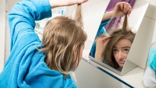 Eine junge Frau betrachtet im Spiegel ihren Haaransatz: Der Teil der Haare, der grau ist, kann nicht plötzlich wieder farbig werden. Aber graue Haare können an der Wurzel wieder farbig weiterwachsen, zumindest für einen gewissen Zeitraum. Zu diesem Ergebnis kam eine Studie im Jahr 2021.