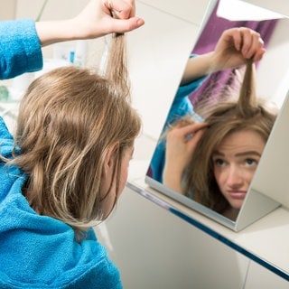 Eine junge Frau betrachtet im Spiegel ihren Haaransatz: Der Teil der Haare, der grau ist, kann nicht plötzlich wieder farbig werden. Aber graue Haare können an der Wurzel wieder farbig weiterwachsen, zumindest für einen gewissen Zeitraum. Zu diesem Ergebnis kam eine Studie im Jahr 2021.