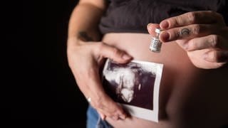 Frau hält Ultraschallbild und Impfampulle vor ihren schwangeren Bauch.
