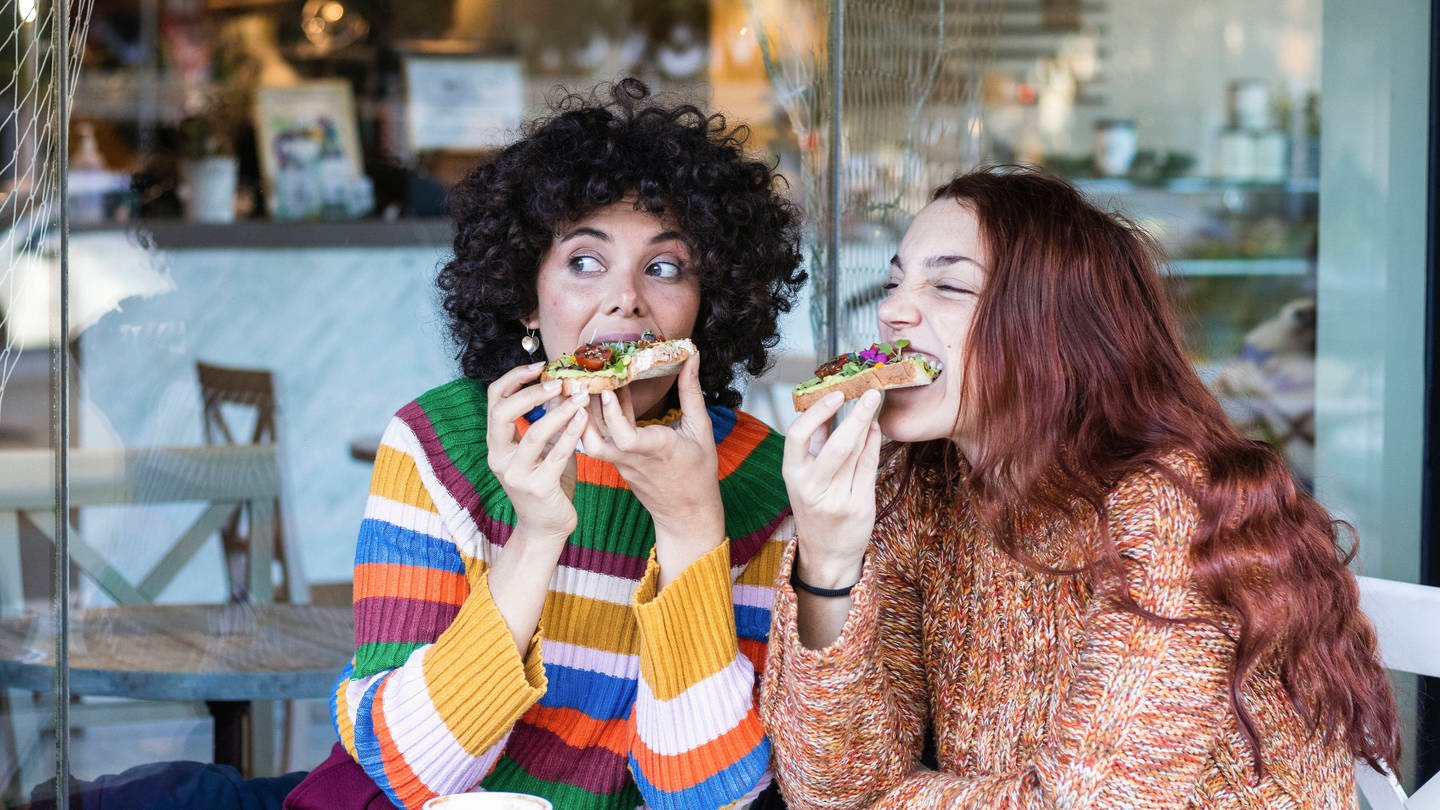 Zwei Frauen beißen beim Frühstück herzhaft in belegte Brote.