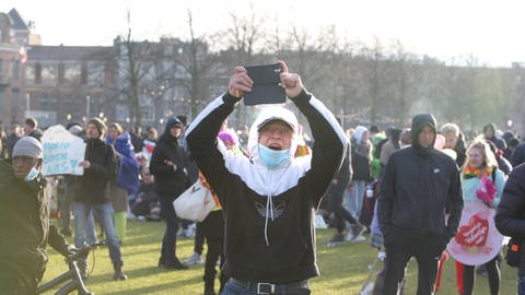 Ein Anti-Lockdown-Demonstrant trägt eine Maske und hält schreiend ein Smartphone nach oben.