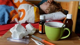 Ein Junge liegt mit Fieber im Bett. Vor ihm auf dem Tisch steht ein Fiebersaft