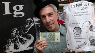 Zeremonienmeister Marc Abrahams hält einen 10-Billionen-Dollar-Schein und den Ig-Nobelpreis 2022 in seinem Büro.