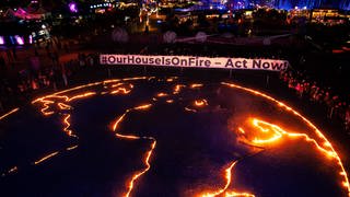 Das Bild zeigt eine Aktion von Fridays for Future in München auf der Theresienwiese. Auf einem Plakat steht: #OurHouseIsOnFire - act now.
