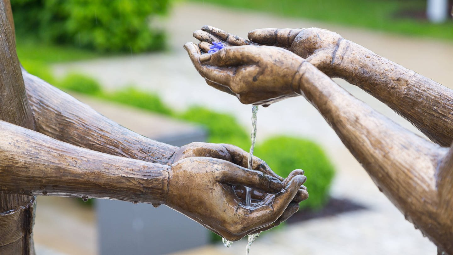 Hände zweier Bronzefiguren, die sich Wasser reichen: Im Mittelalter hat man mit den Fingern gegessen. Zur Reinigung der Hände wurden Wasserschalen gereicht. Allerdings geschah das nicht auf Augenhöhe. Von diesem Brauch leitet sich die Redensart ab: Jemandem nicht das Wasser reichen können