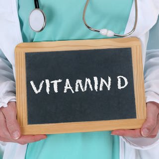 Arzt hält ein Schild mit der Aufschrift "Vitamin D" hoch. Generell sollte Vitamin D nur mit Absprache eines Arztes oder einer Ärztin zu sich genommen werden.