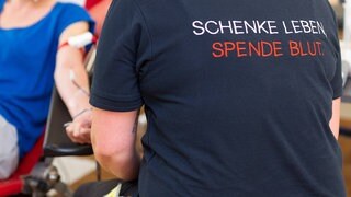 Im Bild sieht man eine Person vom DRK bei der Entnahme vom Blut eines Spenders, auf dem Rücken Ihres T-Shirts steht: Schenke Leben, spende Blut.