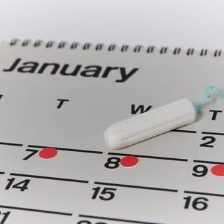 Wandkalender, darauf liegt ein Tampon und mit roten Punkten sind die Tage der Periode im Kalender eingetragen