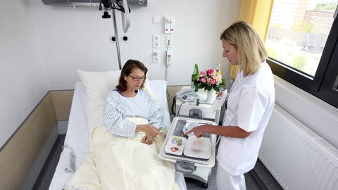  Patientin in einem Einzelzimmer, im Krankenbett. Krankenschwester bringt eine Mahlzeit auf einem Tablett. Symbolbild.