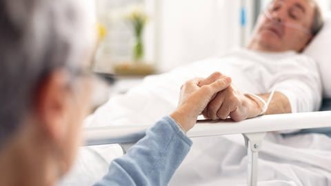 Ein Mann liegt schwer krank in einem Krankenhausbett, seine Frau hält ihm die Hand. Symbolbild.