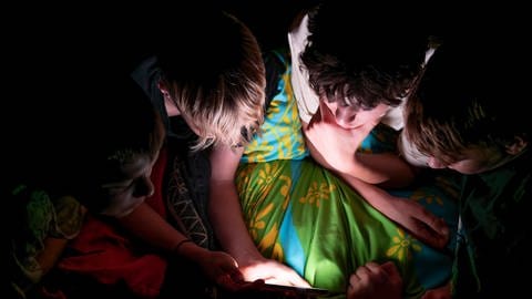 Jungs schauen nachts vor dem Schlaf auf ein Handy mit hellem Bildschirm. Kein empfehlenswerter Tipp. Symbolbild.