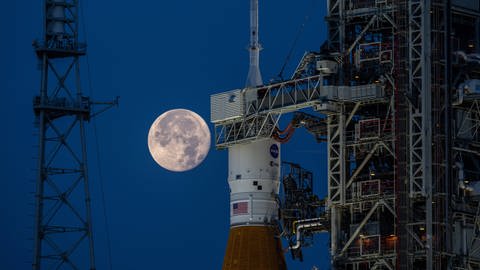 Die Spitze des Space Launch Systems, der Mond-Rakete des Artemis-Programms, mit dem Orion-Raumschiff bei Dämmerung. Im Hintergrund ist der Mond zu sehen.