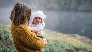 Baby auf dem Arm seiner Mutter vor einem See im Hintergrund