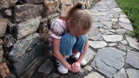 Mädchen hockt vor einer Mauer. Wenn Kinder aufgrund von Rheuma ständig heftige Schmerzen haben, beginnen sie, schmerzauslösende Bewegungen zu vermeiden