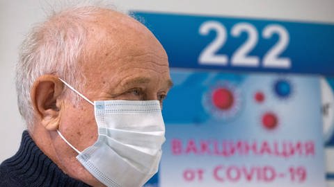 Alter Mann steht mit Maske vor einem blauen Schild mit russischer Covid-19-Beschriftung