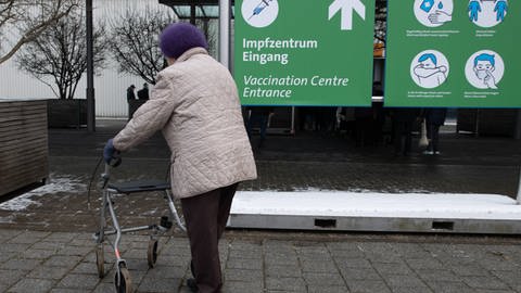 Eine Seniorin mit einem Rollator auf dem Weg zu einem Impfzentrum. Sie trägt eine lilafarbene Mütze und eine beige Jacke und folgt der Beschilderung "Impfzentrum Eingang".