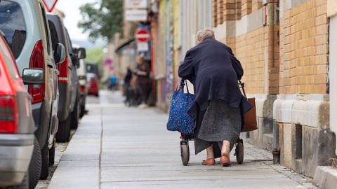 Alte Frau geht gekrümmt am Rollator: Rollatoren sollten nicht langfristig benutzt werden. Denn sie verändern das Gangbild und die Fähigkeit, selbst das Gleichgewicht zu halten, geht verloren.  Das führt im Alltag für mehr Stürzen. Auch die Fitness leidet.