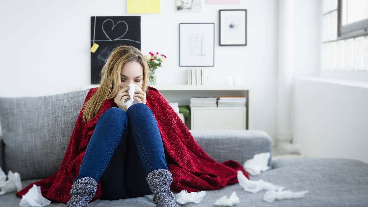 Eine Frau mit einer Erkältung sitzt im Herbst auf einem Sofa. Sie putzt sich die Nase und hat eine Decke über den Schultern liegen, denn im Herbst schlägt die Erkältungswelle wieder zu.