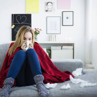 Eine Frau mit einer Erkältung sitzt im Herbst auf einem Sofa. Sie putzt sich die Nase und hat eine Decke über den Schultern liegen, denn im Herbst schlägt die Erkältungswelle wieder zu. 