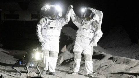 Zwei Astronauten winken in der LUNA-Halle in Köln können in die Kameras. Die LUNA-Halle steht auch für internationale Partner, wie die NASA offen. Hier können sich angehende Astronauten in der Trainingshalle auf künftige Mondmissionen vorbereiten. 