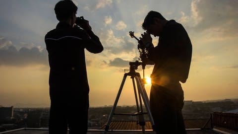 Zwei Personen stehen auf einem Dach, während die Sonne untergeht. Einer der Männer schaut durch ein Fernglas, der andere durch ein Teleskop in den Himmel. Der Komet C2023 A3 Tsuchinshan-Atlas droht auseinander zu brechen.