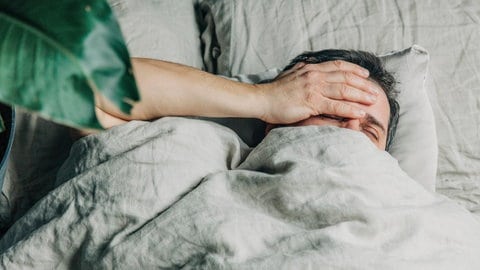 Ein Mann liegt im Bett und hält sich die Hand an die Stirn. Die Zeitumstellung von Normalzeit auf Sommerzeit belastet den Chronotyp Eule besonders. 