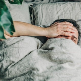 Ein Mann liegt im Bett und hält sich die Hand an die Stirn. Die Zeitumstellung von Normalzeit auf Sommerzeit belastet den Chronotyp Eule besonders. 