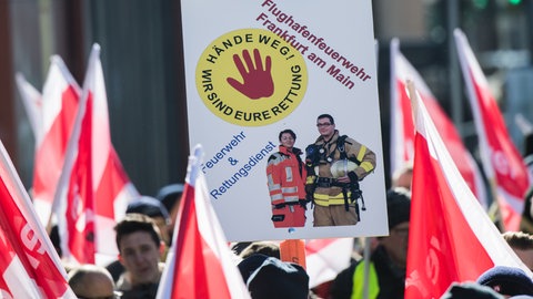 Ein Plakat "Hände weg! Wir sind Eure Rettung" wird 2018 von einem Mitglied der Flughafenfeuerwehr Frankfurt auf einer Demonstration gehalten