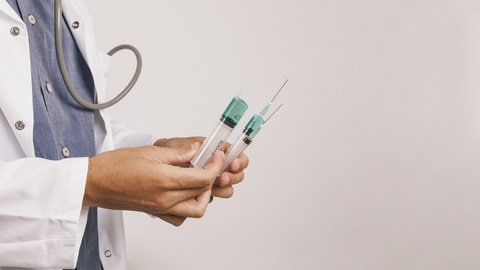 Symbolbild: Ein Arzt, man sieht nur dessen Oberkörper, hält mehrere Spritzen in der Hand. Er steht seitlich vor einer weißen Wand. Notärztinnen geben psychiatrischen Notfällen seltener eine Spritze zur Beruhigung.